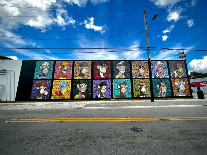 A street-level view of a long outdoor mural composed of multiple colorful panels featuring various cartoon monkey characters, set against a bright blue sky with scattered clouds.