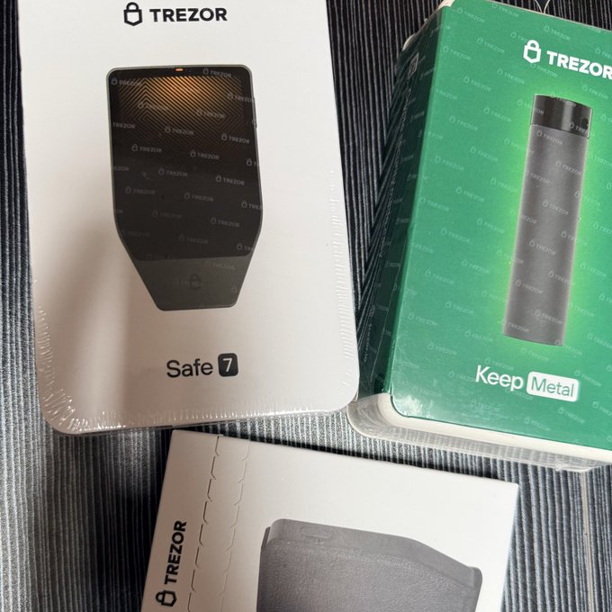 Three Trezor products are displayed on a striped surface: a sealed 'Safe 7' hardware wallet box on the left, a green 'Keep Metal' package on the right, and another Trezor box partially visible at the bottom.