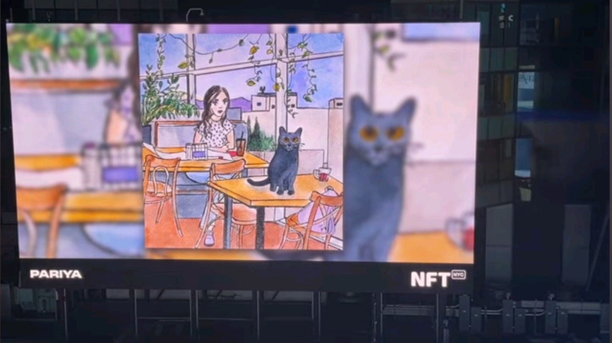 A large NFT NYC billboard featuring the artwork 'PARIYA', depicting an illustrated girl sitting at a café table with a black cat, with a magnified cat image on the right.