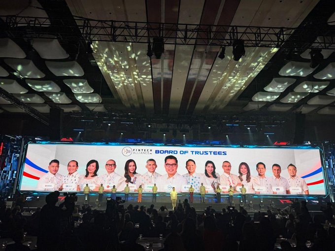 A photograph of a large event stage with a screen displaying 'FINTECH ALLIANCE PH BOARD OF TRUSTEES' and photos of board members, taken from the audience perspective.