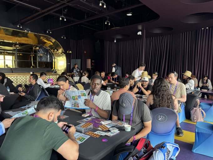 A bustling crowd of attendees sits at long tables in the Purple Room, deeply engaged with cards, trays of tokens, and reference materials for the Pudgy Penguins Vibes Trading Card Game event presented by @PudgyCanada.