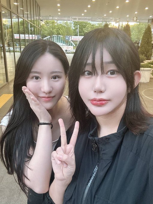 A selfie of two young East Asian women posing together outdoors; one is making a peace sign while the other holds her hand to her cheek.