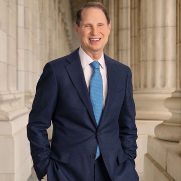 Professional portrait of a smiling man in a navy blue suit with a light blue tie, standing between classical white columns.