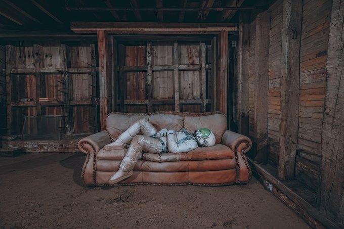 An astronaut in a spacesuit reclining on a brown leather couch inside an abandoned, weathered wooden barn.