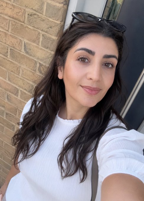 A selfie of a woman with long dark hair wearing a white ribbed top, with sunglasses resting on her head, standing outdoors in front of a brick wall.