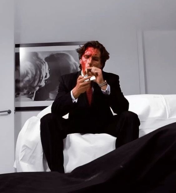 A man in a black suit sits on a white chair wiping blood from his face with a handkerchief, with a black-and-white photograph visible on the wall behind him.
