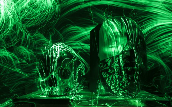 A green-tinted, abstract image with swirling light trails forming what appears to be a hooded figure and a skeletal or wireframe shape.