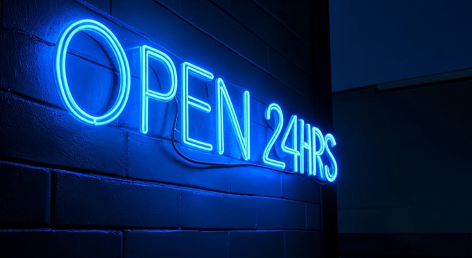 A blue neon sign reading 'OPEN 24HRS' glows brightly against a dark brick wall at night.