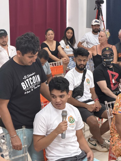 A young man speaks into a microphone while wearing a "Bitcoin Berlin" t-shirt, surrounded by attendees at what appears to be a Bitcoin community event, with others wearing apparel referencing "El Salvador" and "Bitcoin Beach," aligning with the post's mention of the Bitcoin Berlin SV movement and its local impact in El Salvador.