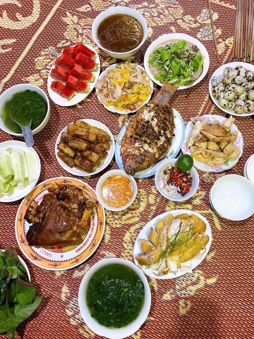 A traditional meal is spread out on a woven mat, featuring a whole fried fish, sliced watermelon, boiled chicken, quail eggs, spring rolls, and various vegetables and dipping sauces.