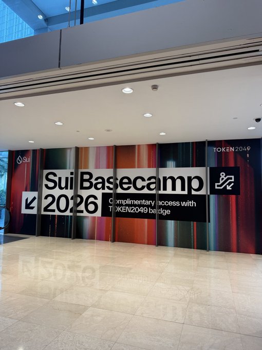 A conference signage for 'Sui Basecamp 2026' promoting complimentary access with a TOKEN2049 badge, displayed alongside colorful vertical banners at an event venue entrance.