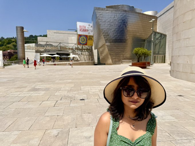The image shows a woman wearing sunglasses and a hat standing outside of a modern building with a glass facade. The woman appears to be posing for a photo while wearing her hat. There are several other people visible in the background, but they do not seem to be the main focus of the photograph. The post includes some text that reads "103F (40C) in Spain. No AC with the Europoors. If only there were a Buc-ee's or a Walmart here. Without ranch sauce, we might not survive." This suggests that the woman is in a foreign country where the weather may be warm and sunny but lacks essential amenities like an air conditioner and grocery stores.