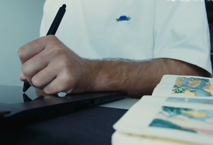 A man in a white t-shirt using a stylus on a graphics tablet, with small watercolor paintings visible on paper beside him.