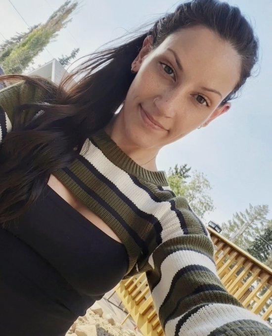 A selfie of a smiling woman with long dark hair, wearing a green and white striped cardigan over a black top, taken outdoors with trees and a wooden structure visible in the background.