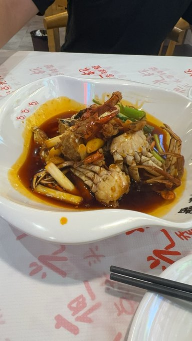 A plate of seafood—appearing to include crab and lobster pieces—in a dark savory sauce with green onions, served at a restaurant table with Chinese characters visible on the placemat.