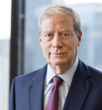 A professional headshot of an older man with gray hair wearing a navy blue suit jacket, light blue shirt, and burgundy tie, photographed indoors with a blurred city window background.