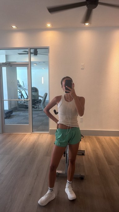 A woman in a white tank top, green shorts, and white sneakers takes a mirror selfie with wired earbuds in her phone while standing on a wood floor with gym equipment visible through a glass door behind her. The post connects her post-workout moment with the parallel act of securing Bitcoin savings ("sats") in cold storage, framing both running and crypto custody as disciplined self-care routines.