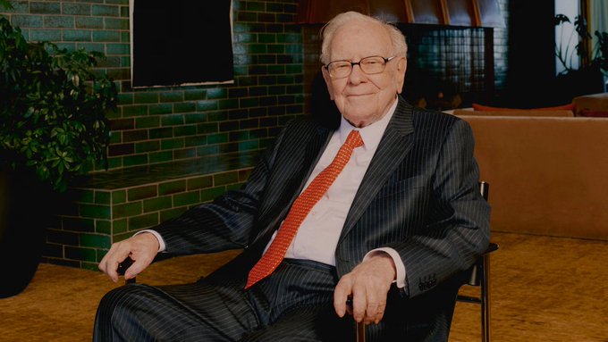 A portrait of Warren Buffett seated in a chair, wearing a dark pinstripe suit with a red tie, smiling against a green tiled wall background.
