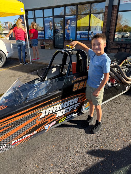 A young boy poses proudly next to a sleek black dragster labeled "JARMAN NEFF" with the number "588" and sponsor logos visible, suggesting he got an up-close look at a junior drag racing car at a local event. In the background, two women chat under a yellow canopy tent, tying into the post's theme of supporting small businesses and enjoying a casual day outdoors at what appears to be a racing community gathering.