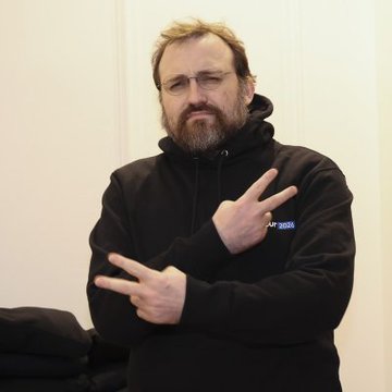 A bearded man with glasses, wearing a black hoodie with "GAMP 360" text visible, makes a peace sign with both hands crossed in front of his chest, appearing to make a defiant gesture that aligns with Charles Hoskinson's strong stance against Senator Warren's crypto policies.