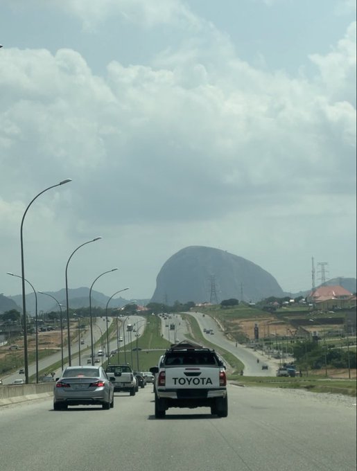 A view from a vehicle on a multi-lane highway showing cars ahead, including a white Toyota pickup truck, with a large rocky mountain visible in the distance under a cloudy sky.