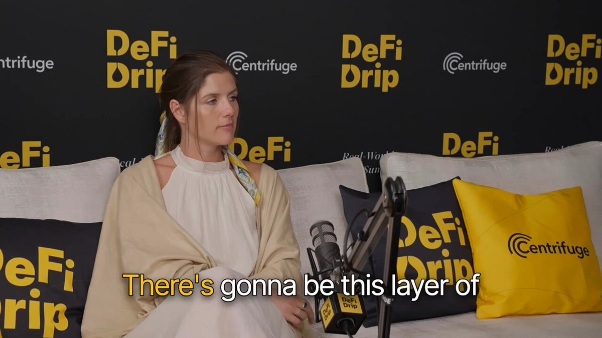 The image features a woman sitting on a couch with her legs crossed while holding a microphone. The background of the image includes a black wall with yellow text that reads "Defi" and "DIP". There are also two pillows visible, one behind the woman and another further to the right side of the frame.
