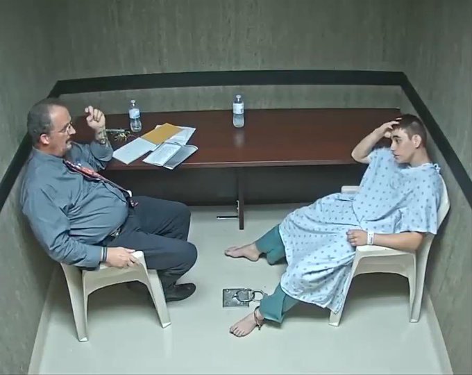An overhead view shows a man in a suit and tie sitting across a small table from a barefoot person wearing a hospital gown, appearing to be in an interview room.