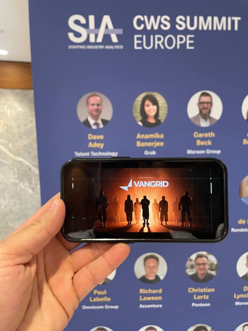 A hand holding a smartphone displaying the 'VANGRID' app in front of a 'CWS SUMMIT EUROPE' banner featuring various speaker headshots including Dave Adey, Anamika Banerjee, and Gareth Beck.