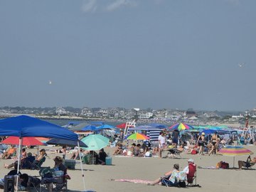 A photograph of a crowded beach scene with people sitting under colorful umbrellas on the sand, with the ocean and a coastline visible in the background under a hazy sky.