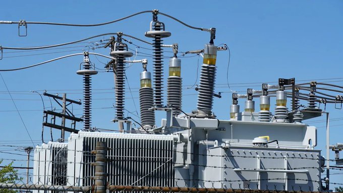 An outdoor view of an electrical power substation featuring large transformers, ceramic insulators, and high-voltage equipment under a clear blue sky.