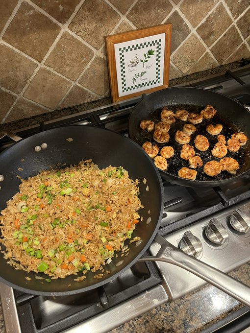 Two pans cooking on a gas stove—one with fried rice mixed with vegetables and the other with browned shrimp or chicken pieces, with a framed picture in the background.