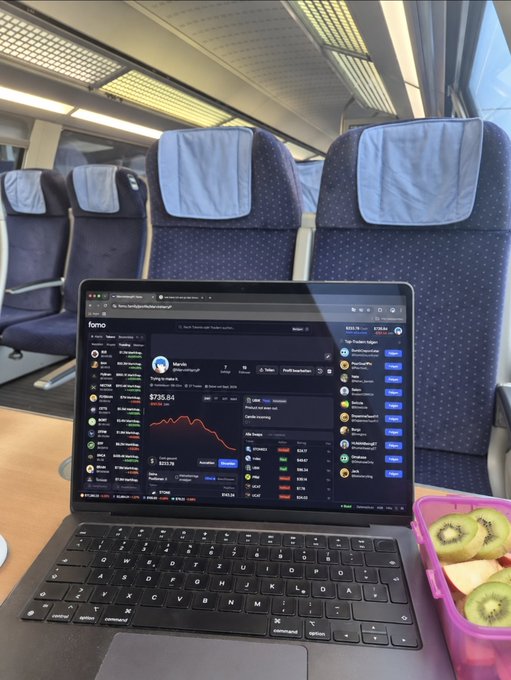 A laptop on a train displays the Fomo crypto trading app with an Arbitrum chart showing $735.84, next to a pink lunchbox containing kiwi slices and other snacks.