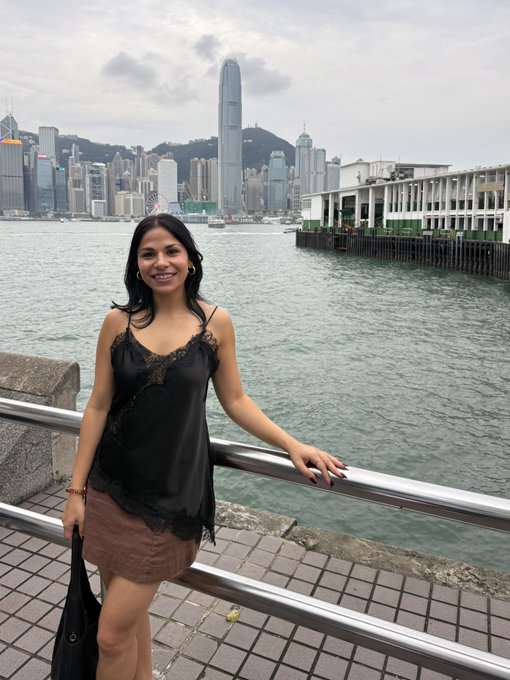 A woman smiles while posing in front of the Hong Kong skyline. The accompanying post "see ya later HK!" indicates she is leaving Hong Kong.
