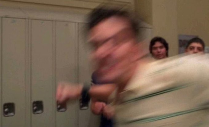 A blurry photograph of a man in a striped shirt standing in front of school lockers, seemingly in motion, with two other people visible in the background.