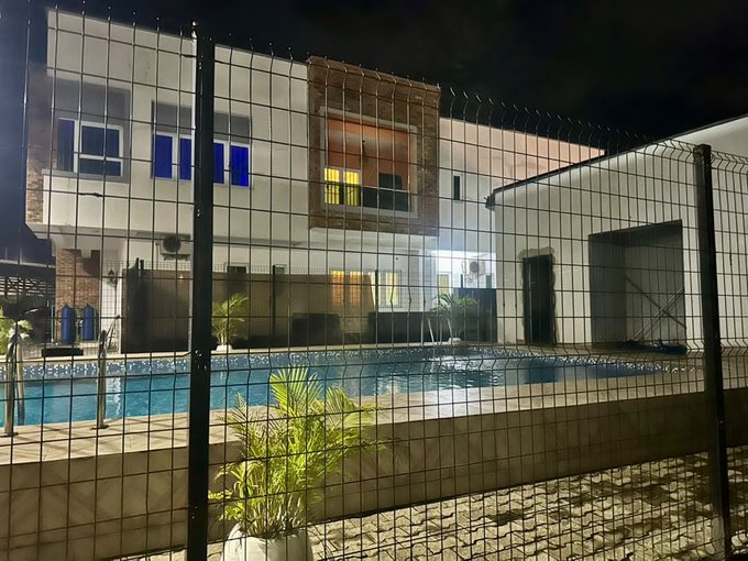 A nighttime view of a modern two-story building with blue-lit windows and a swimming pool, seen through a wire mesh fence.