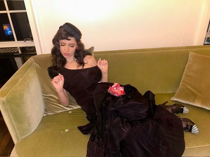A woman wearing a black off-shoulder dress and black beret lounges on a green velvet couch, holding what appears to be a dark object, with a red item resting on her lap.