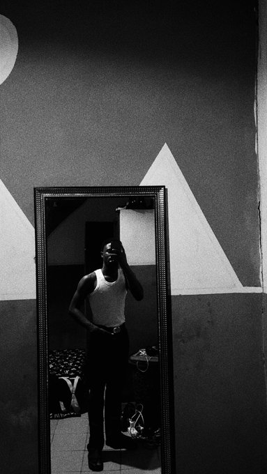 A black-and-white photo shows a man in a white tank top and dark pants standing in front of a tall mirror, taking a picture with his phone in a room featuring geometric wall designs.
