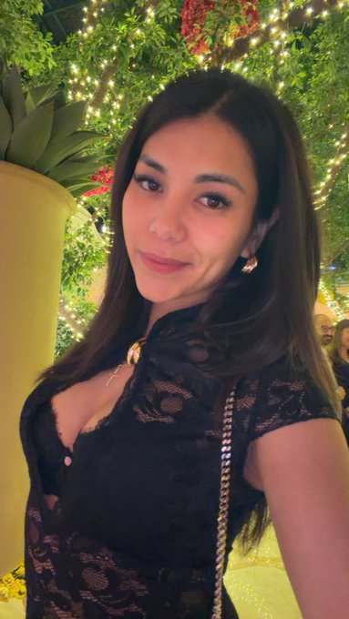A selfie of a young woman with long dark hair wearing a black lace top and gold jewelry, posed in front of greenery decorated with string lights.