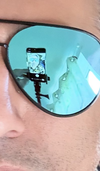A close-up of a person's face shows blue mirrored aviator sunglasses reflecting a smartphone mounted on a tripod displaying a camera feed.