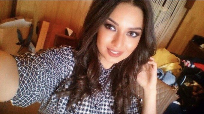 Selfie of a young woman with long dark hair wearing a black and white geometric patterned dress, in a cluttered room setting.