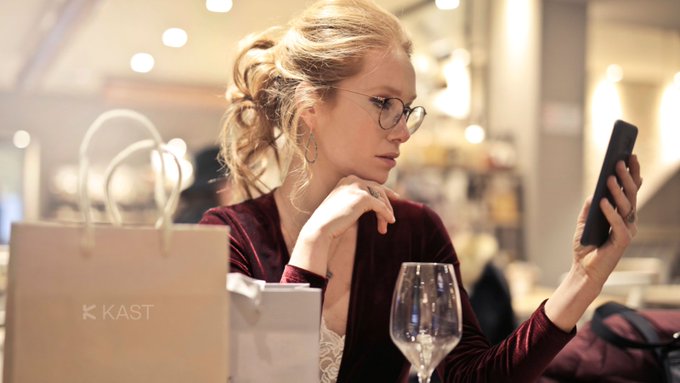 The image shows a woman sitting at a table with her cell phone, looking at something on the screen. She has a wine glass next to her and a shopping bag nearby. The scene takes place in an indoor setting, possibly a restaurant or cafe.
