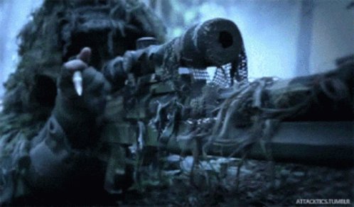 The image shows a person wearing a camouflage outfit holding a gun with a chain attached to it. The person's hand is pointing towards the camera while they are standing next to a wall or a building, possibly at an indoor location like a store or a military base. The caption of the image reads "Ansem shares his ultimate buying guide for $HYPE, $VVV, and $ZEC".