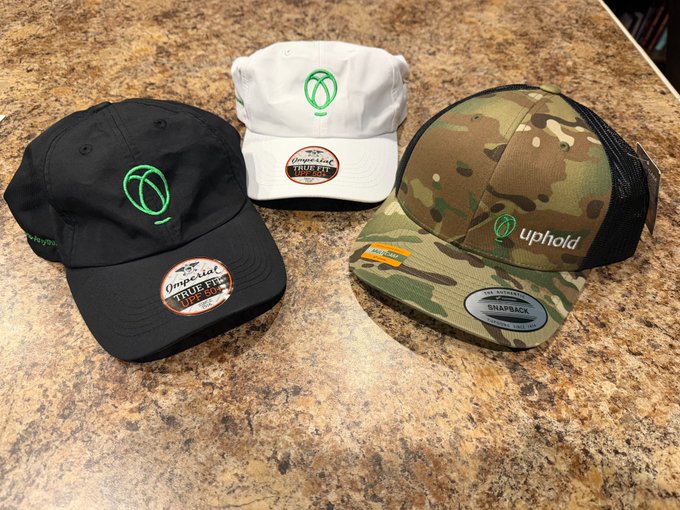 Three baseball caps rest on a granite countertop—two "Imperial TrueFit" caps in black and white, each embroidered with the green Uphold logo, and a camo snapback branded with the Uphold name and logo on the side—appearing to be giveaway prizes for the September 4th @UpholdInc drawing.