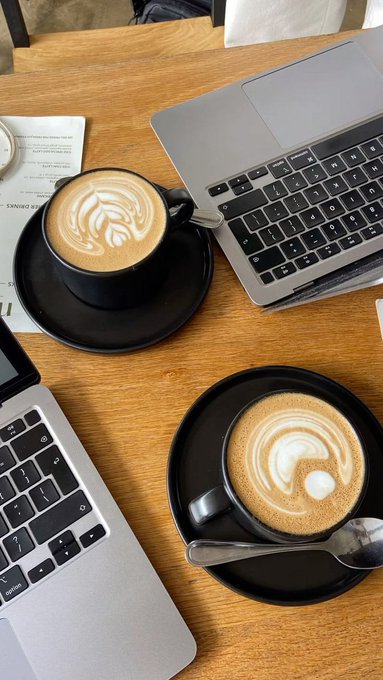 Two cups of latte art coffee sit on a wooden table alongside two open MacBook laptops, capturing a quiet morning work session. The scene reflects the post's "coffee first, then check the market" mindset—a peaceful setup for traders prioritizing a clear head over the trading timeline this Saturday.