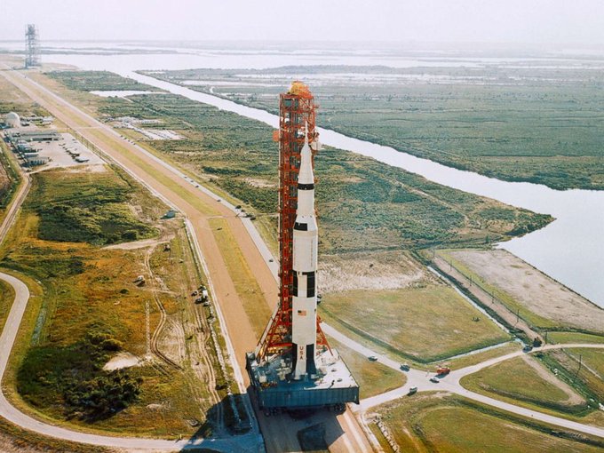 An aerial photograph of a Saturn V rocket on its launch pad at NASA's Kennedy Space Center, surrounded by the launch tower, with coastal waterways and green landscape in the background.
