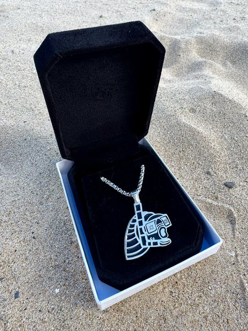 A silver pendant necklace with an Aztec/Mayan warrior design displayed in an open black velvet box on sandy ground.