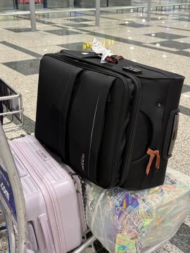 A community manager's loaded luggage cart sits at an airport, featuring a large black suitcase, a pale pink bag, and a plastic-wrapped bundle of product merchandise—visually capturing the heavy physical labor behind their dedication to connecting with NeoSoul builders.