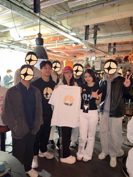 Four friends pose together indoors, with one holding up a white T-shirt featuring a graphic design, their faces obscured by crosshair emojis.