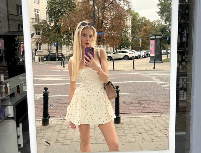 A young blonde woman in a cream polka-dot dress takes a mirror selfie with an iPhone on a city sidewalk, holding a beige handbag with sunglasses perched on her head.