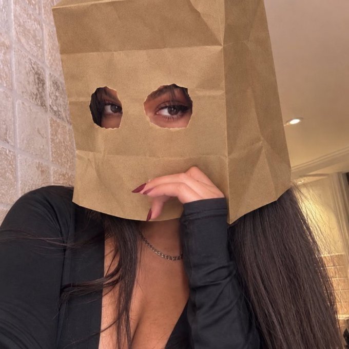 A woman poses playfully with a brown paper bag over her head, which has two eye holes cut out revealing her dark, made-up eyes, while her manicured hand rests beneath the bag and her long dark hair flows down over her black top, humorously illustrating the post's caption "the bag STAYS on $ZCAT" by suggesting she literally cannot remove it.